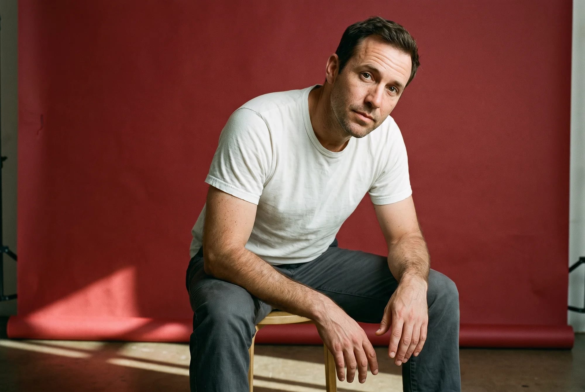 A man sitting on a low stool against oxblood seamless paper, forearms on his knees, looking into the lens.