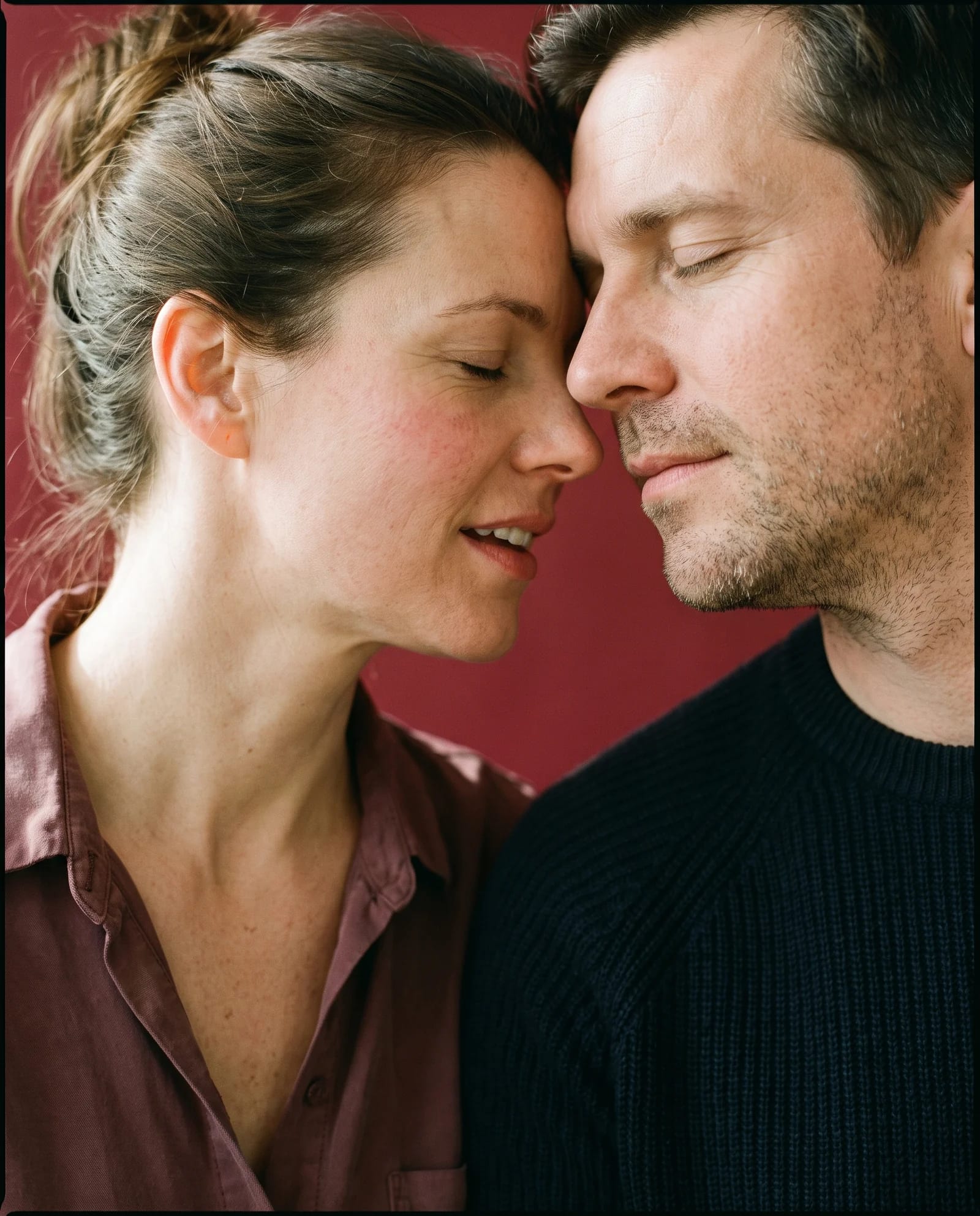 A couple at intimate distance against oxblood seamless paper, her forehead resting against his cheekbone, both with their eyes closed.