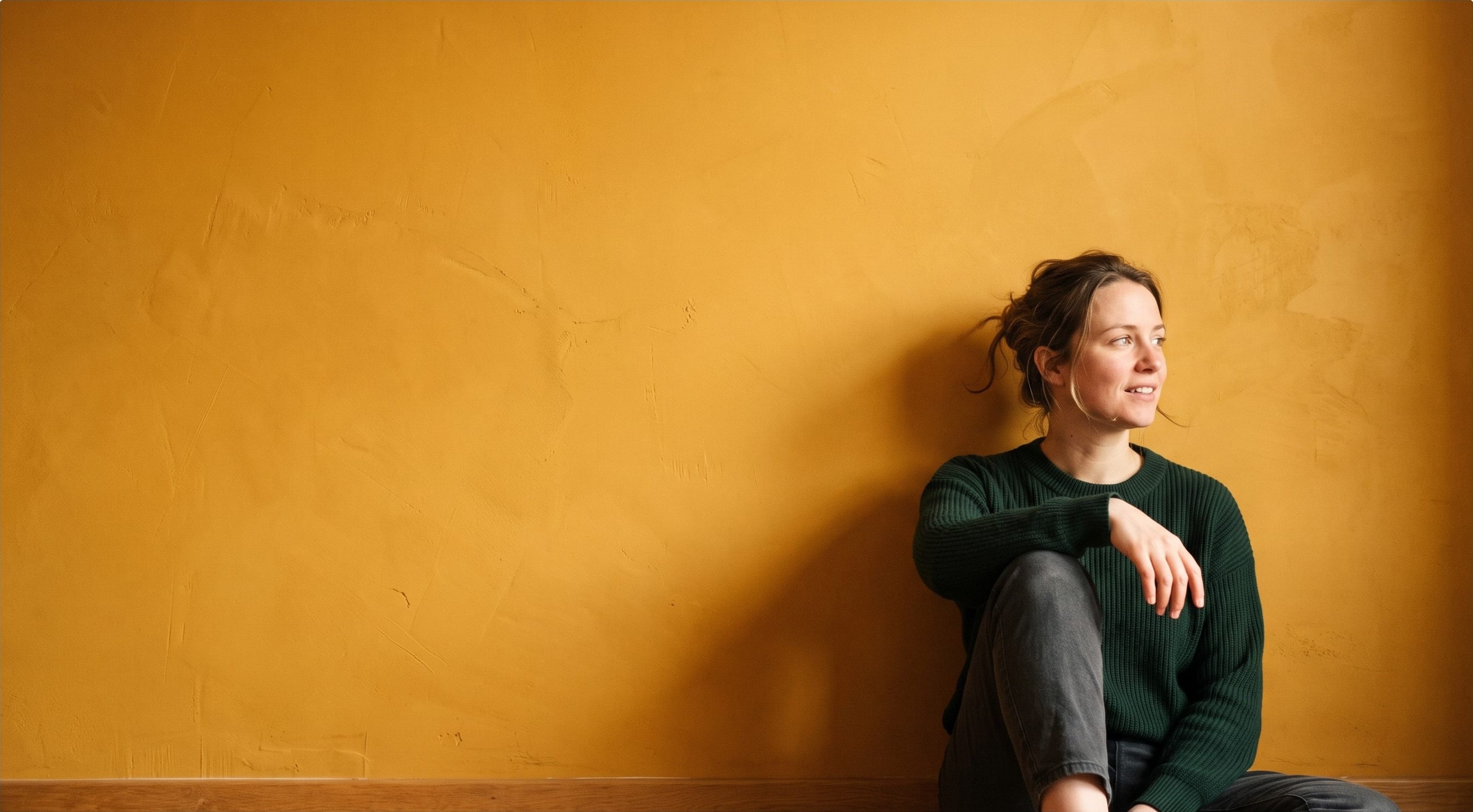 A woman sitting against an amber plaster wall, looking off to one side.