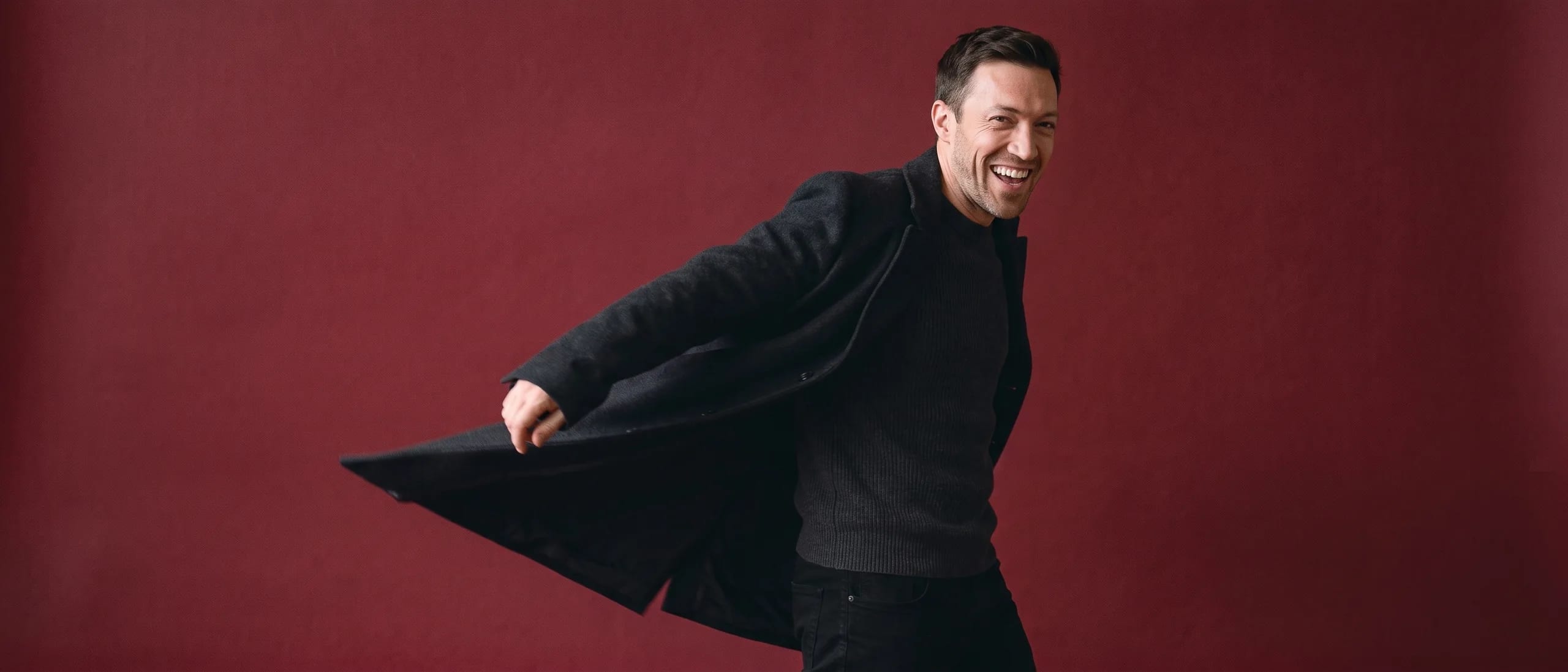 A man shrugging a coat on as he walks out of frame, looking back over his shoulder, laughing