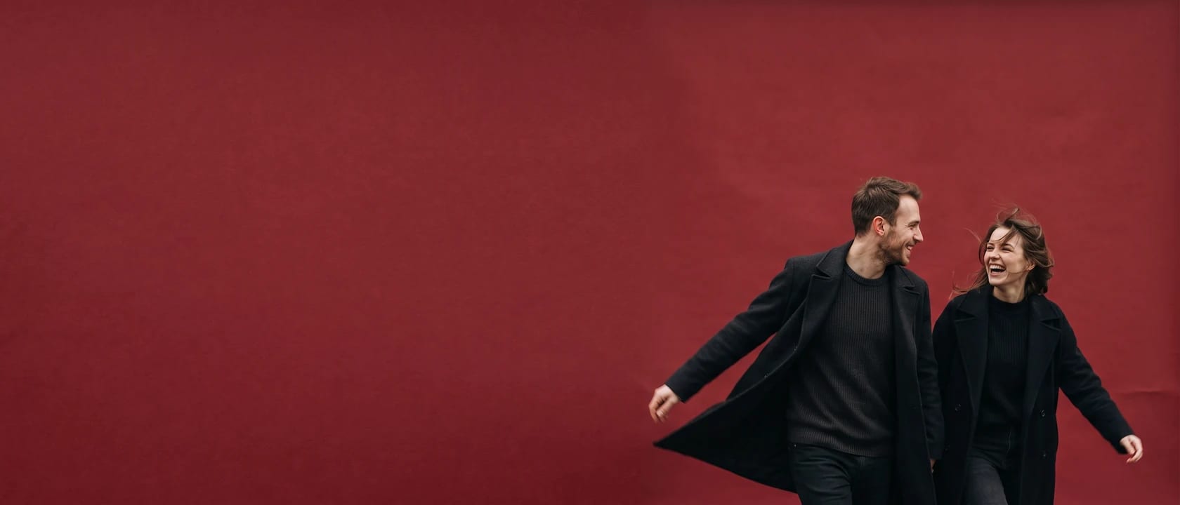 Two people walking fast through the frame together, one turning back mid-laugh, low and right against a wide empty field of oxblood paper