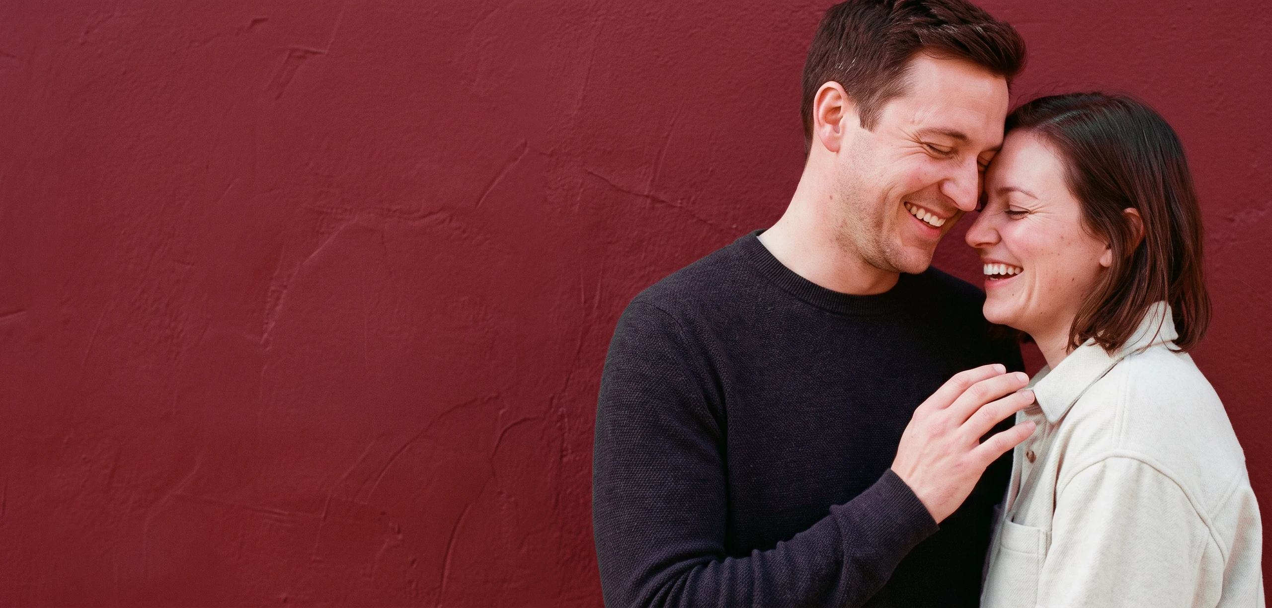 Two people laughing close together against a deep oxblood wall