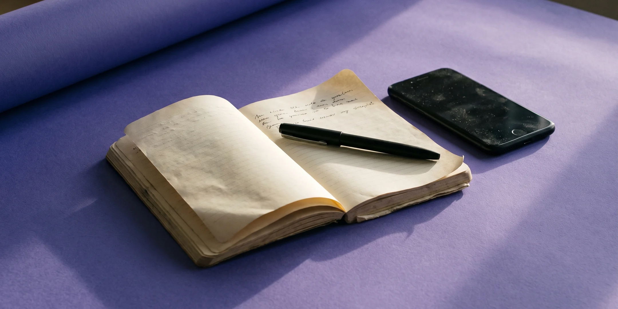An open notebook, a pen and a phone face-down on iris paper in low raking light