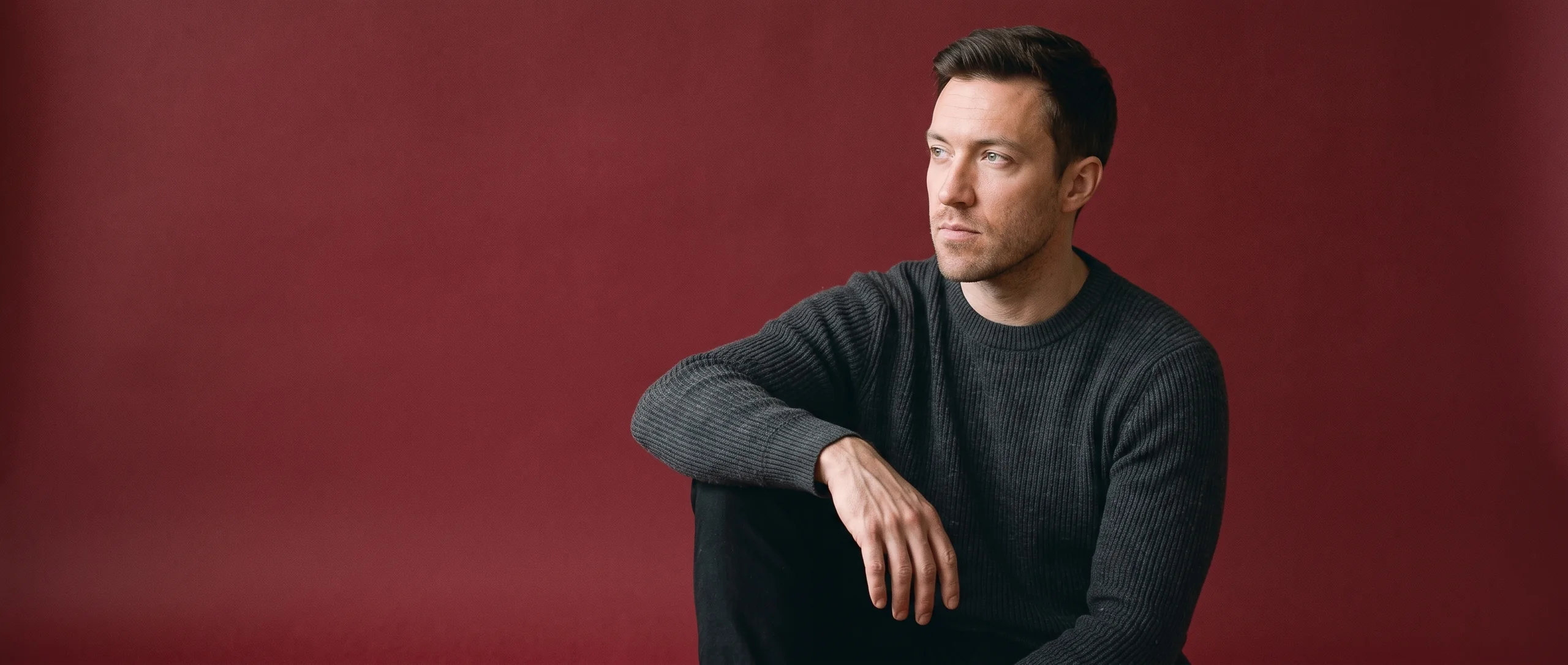 A man sitting on oxblood seamless paper, looking off to one side, alone in a wide field of colour