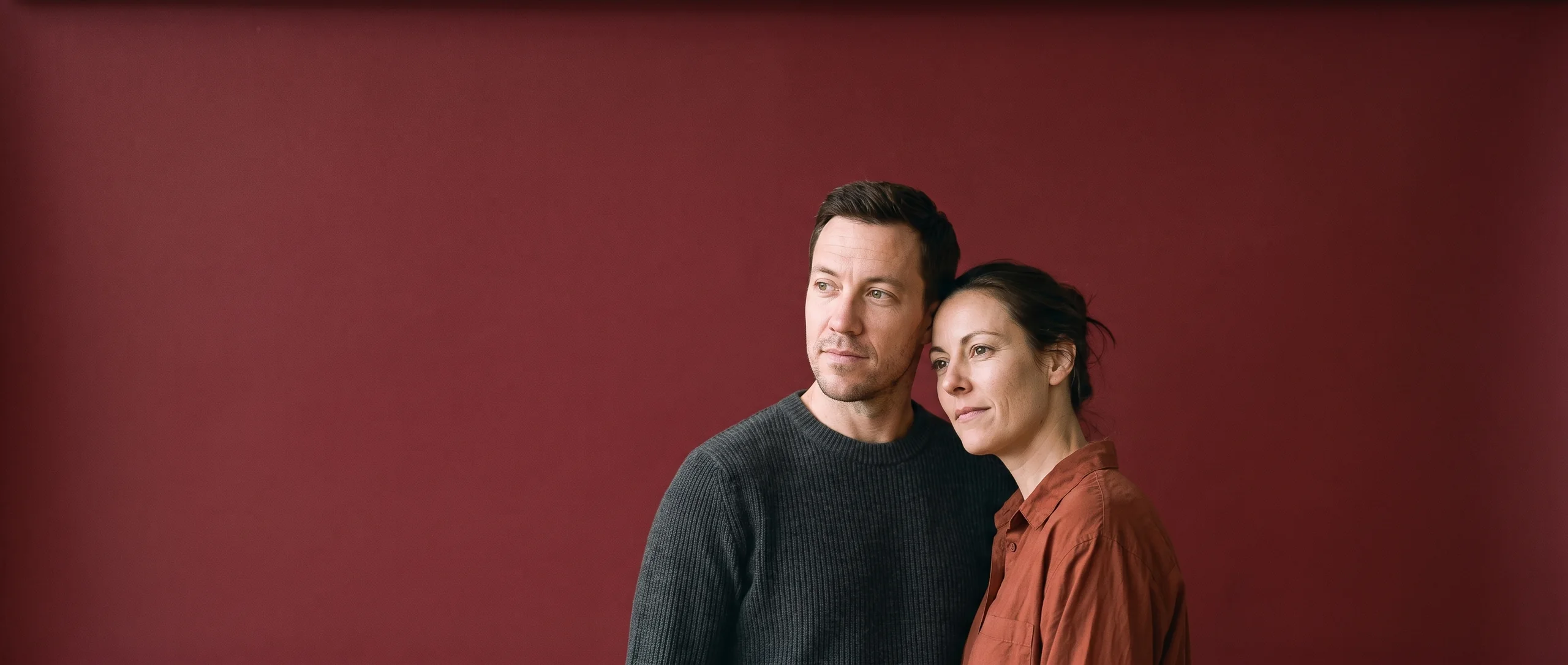 Two people standing close on oxblood seamless paper, looking off to one side