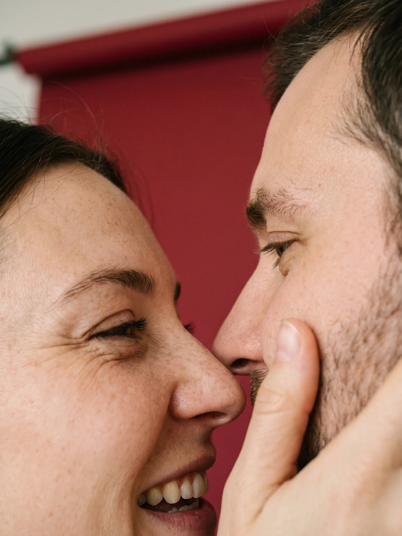 A couple photographed from inches away, the moment before a kiss, against oxblood seamless paper