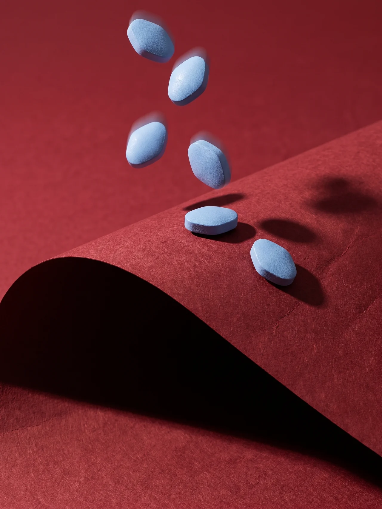 Plain blue sildenafil tablets caught mid-tumble above a fold of oxblood seamless paper