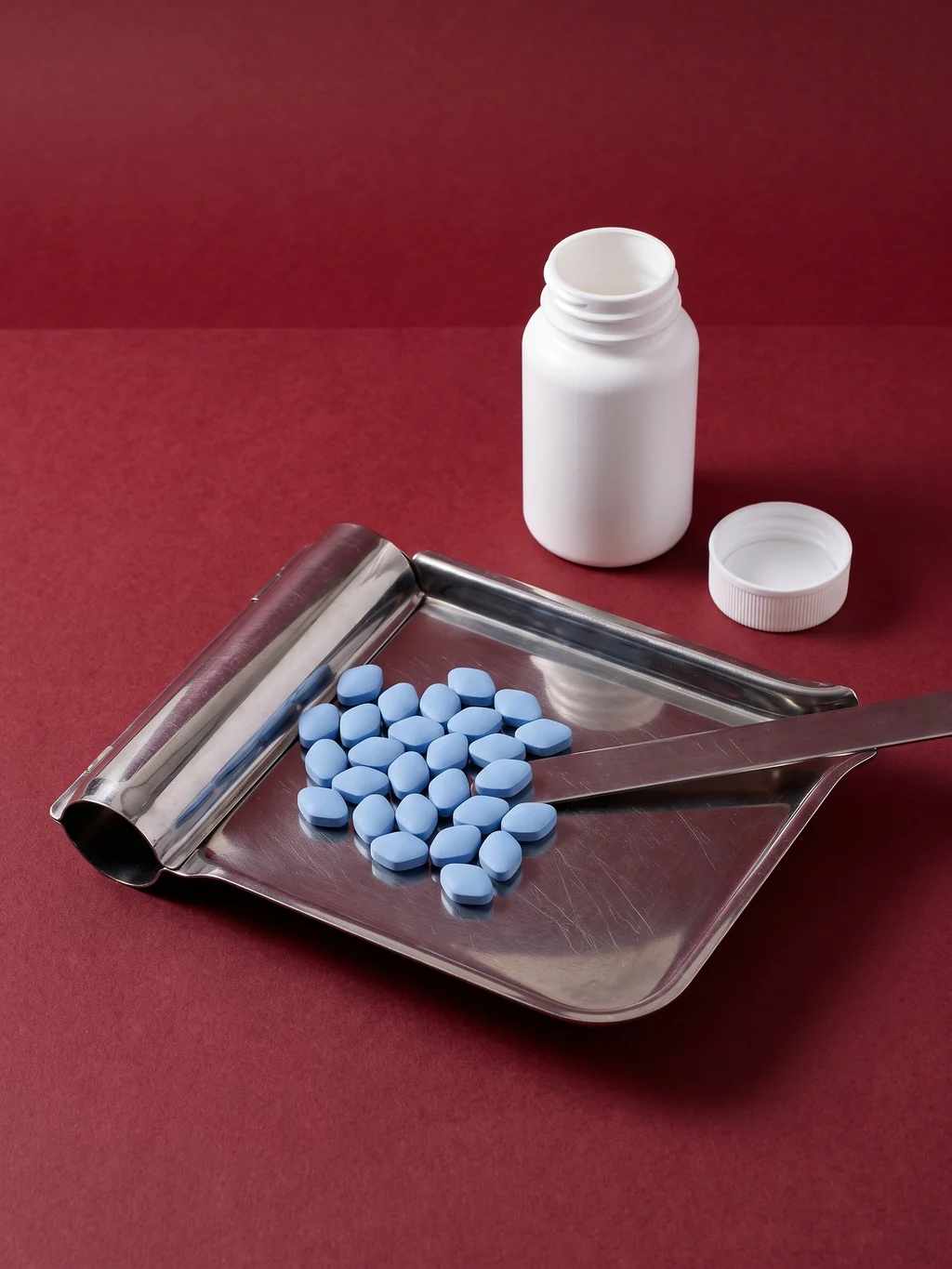 A stainless counting tray and spatula with unmarked tablets gathered mid-count, beside an open unlabelled bottle on oxblood paper