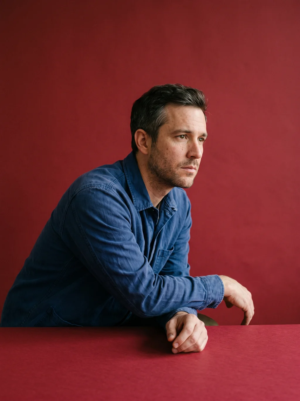 A man seated against oxblood paper, leaning forward, caught mid-thought with nothing in his hands