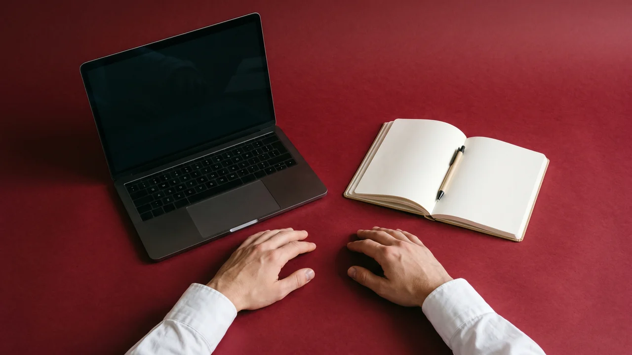 A closed laptop and an open blank notebook on oxblood paper, two hands resting either side