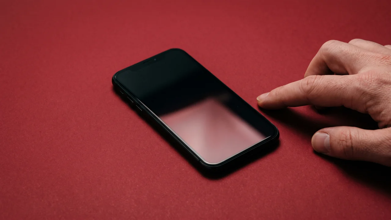 A phone lying face up on oxblood paper, screen dark, a hand resting beside it