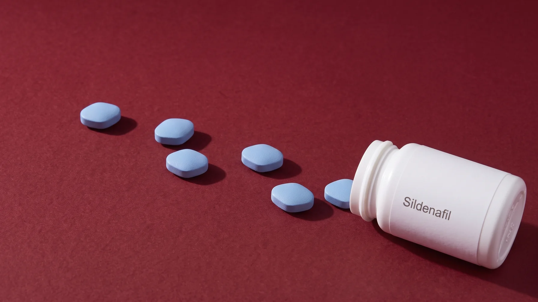 A plain white bottle labelled Sildenafil lying on oxblood paper, tablets running out of it in a diagonal