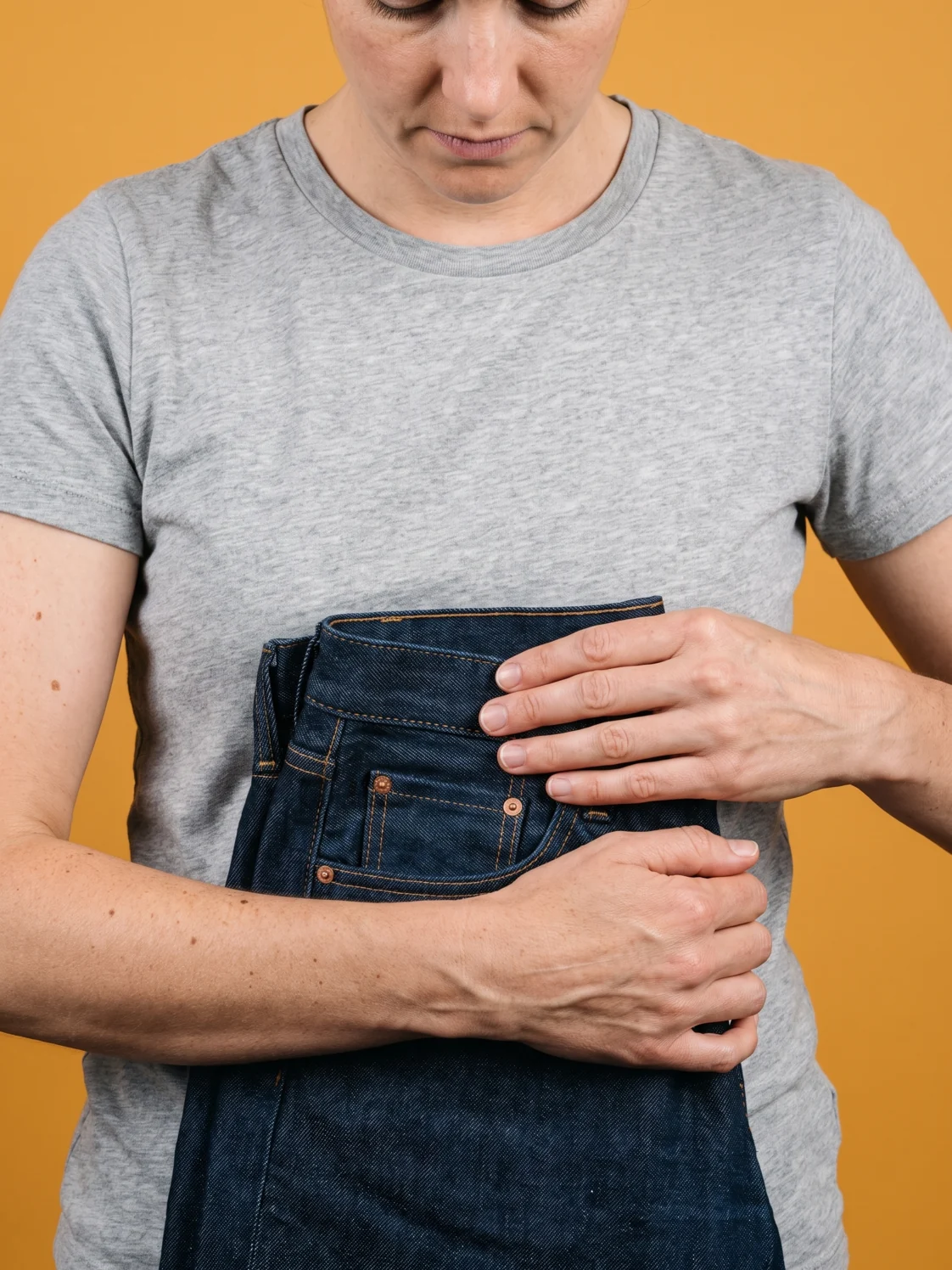 A woman holding a folded pair of jeans flat against her waist, against flat amber seamless paper
